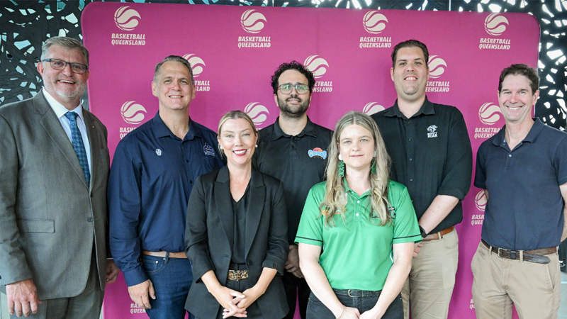 Group of six adults posing for a photo in front of a pink backdrop with Basketball Queensland logos; some wear polo shirts and business attire.