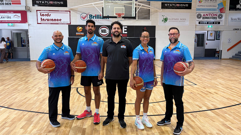 Five people stand on a basketball court posing for a team photo: four players in blue gradient jerseys and two holding basketballs, plus a coach in a black polo, with banners on the wall behind them.