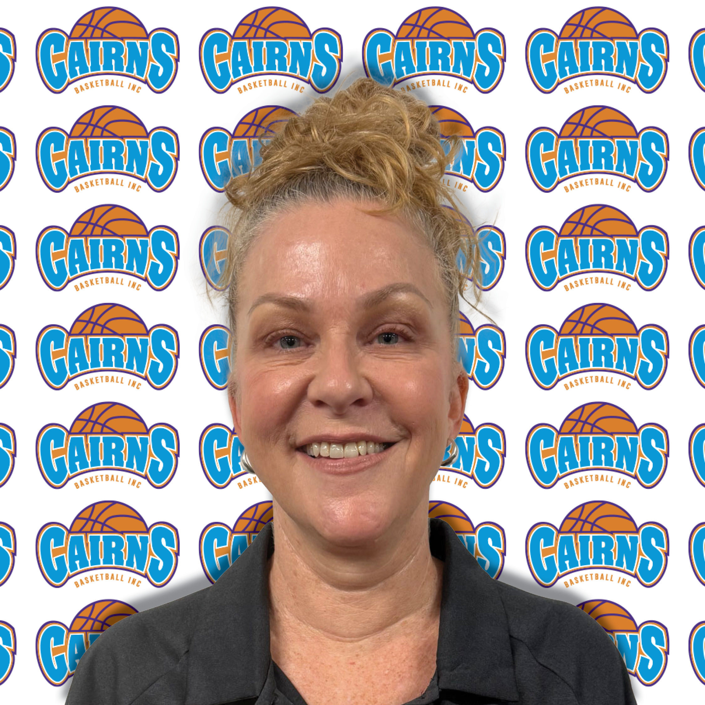 Close-up of a smiling woman with light skin and blonde hair in a bun, wearing a dark blazer, in front of a Cairns Basketball backdrop.