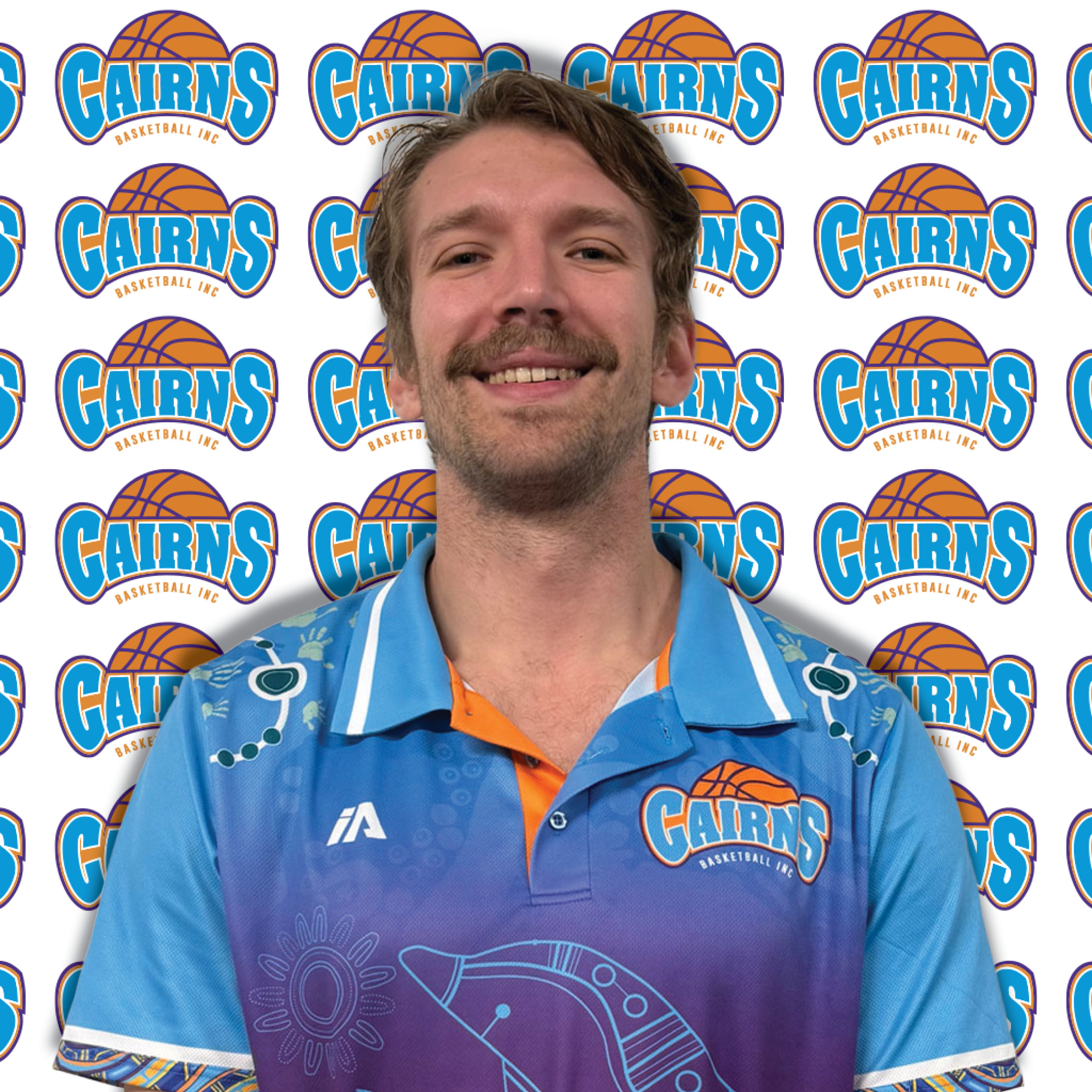 Smiling man in a blue shirt stands in front of a repeating Cairns Basketball Inc. logo backdrop.