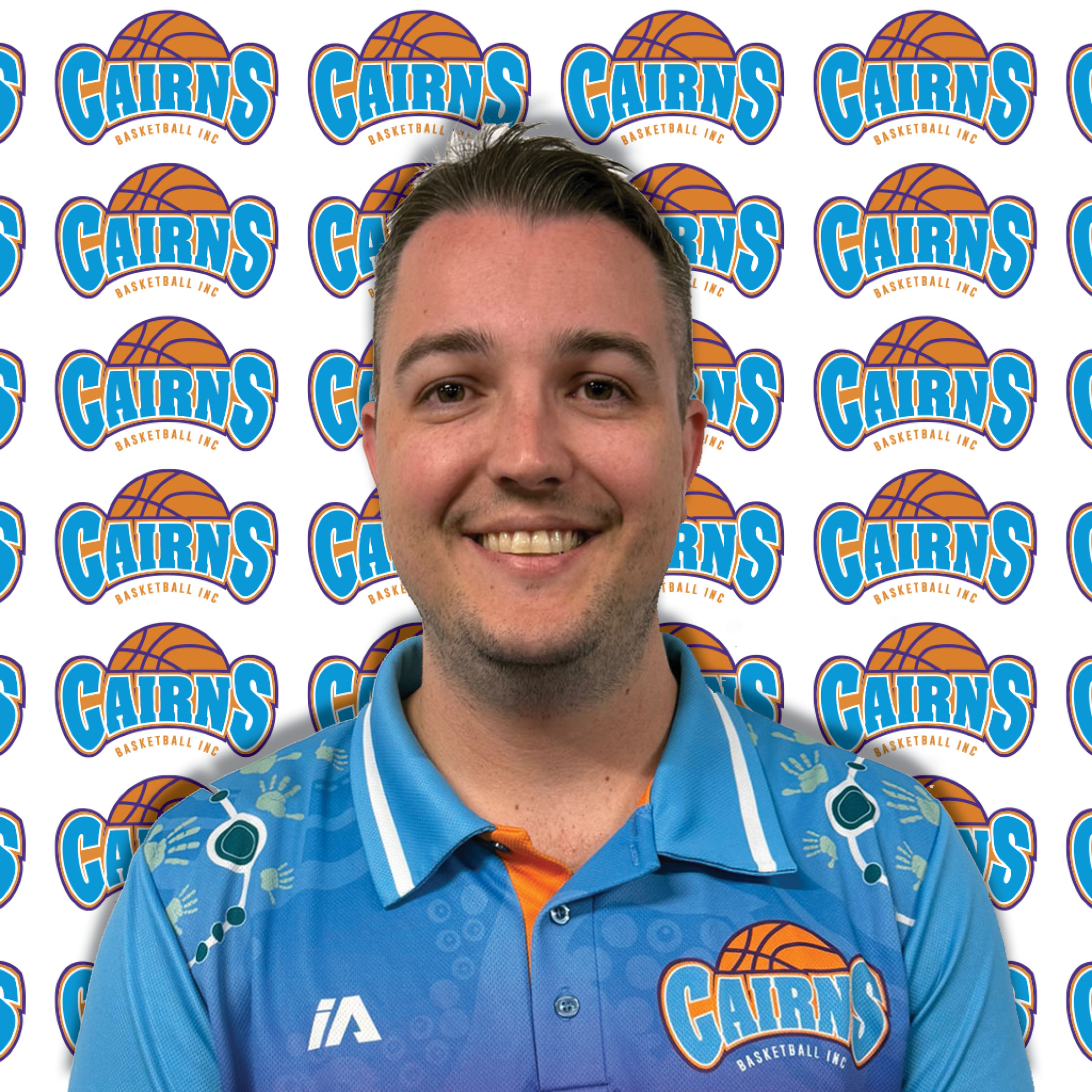 Close-up of a smiling man in a blue basketball jersey in front of Cairns Basketball backdrop logos.