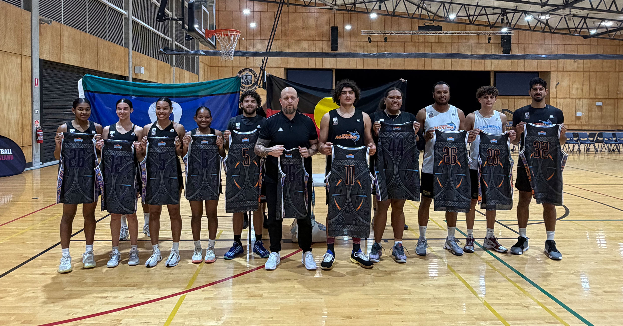 Group of female and male basketball players with a coach posing in a gym, each holding a dark patterned jersey in front of team flags behind them.