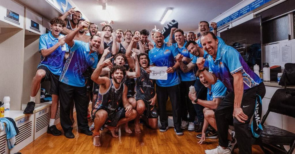 Group of athletes in blue uniforms celebrate a win in a locker room, holding a sign reading 16-0, fists raised.