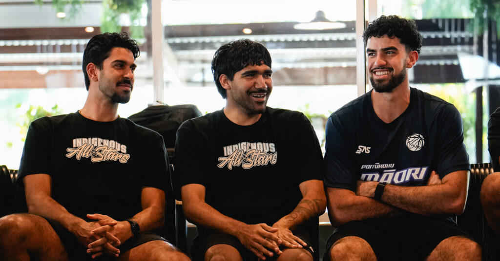 Three men wearing Indigenous All Stars shirts sit together, smiling and chatting indoors.