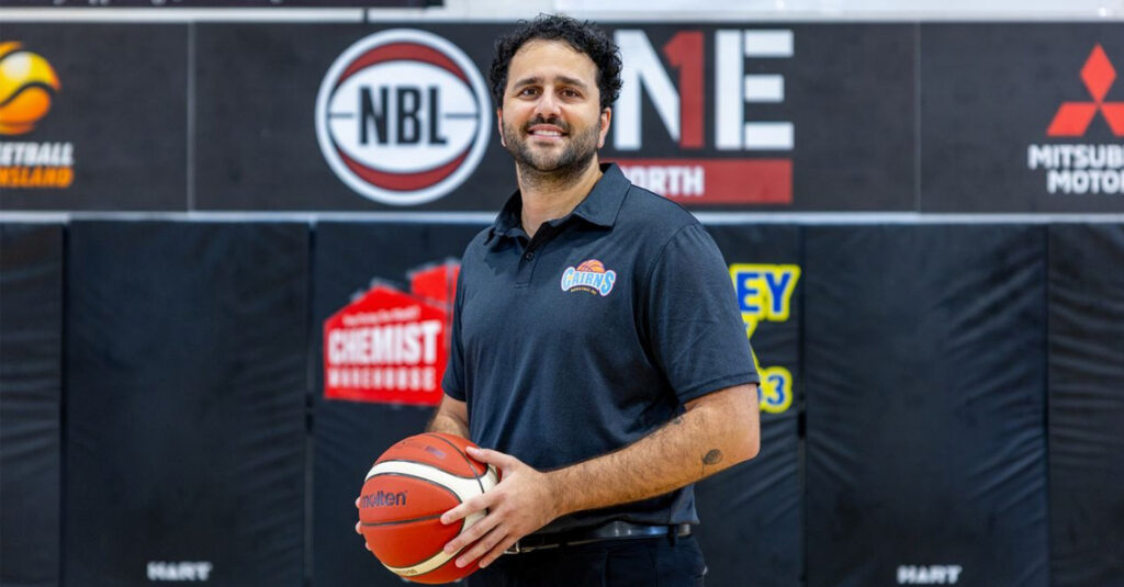 Man in a navy Cairns polo holding a basketball inside a gym with sponsor banners behind him.