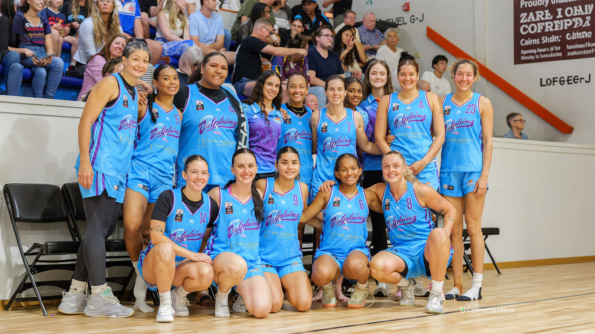 Girls basketball team in blue uniforms posing for a group photo in a gym with spectators in the bleachers behind them.